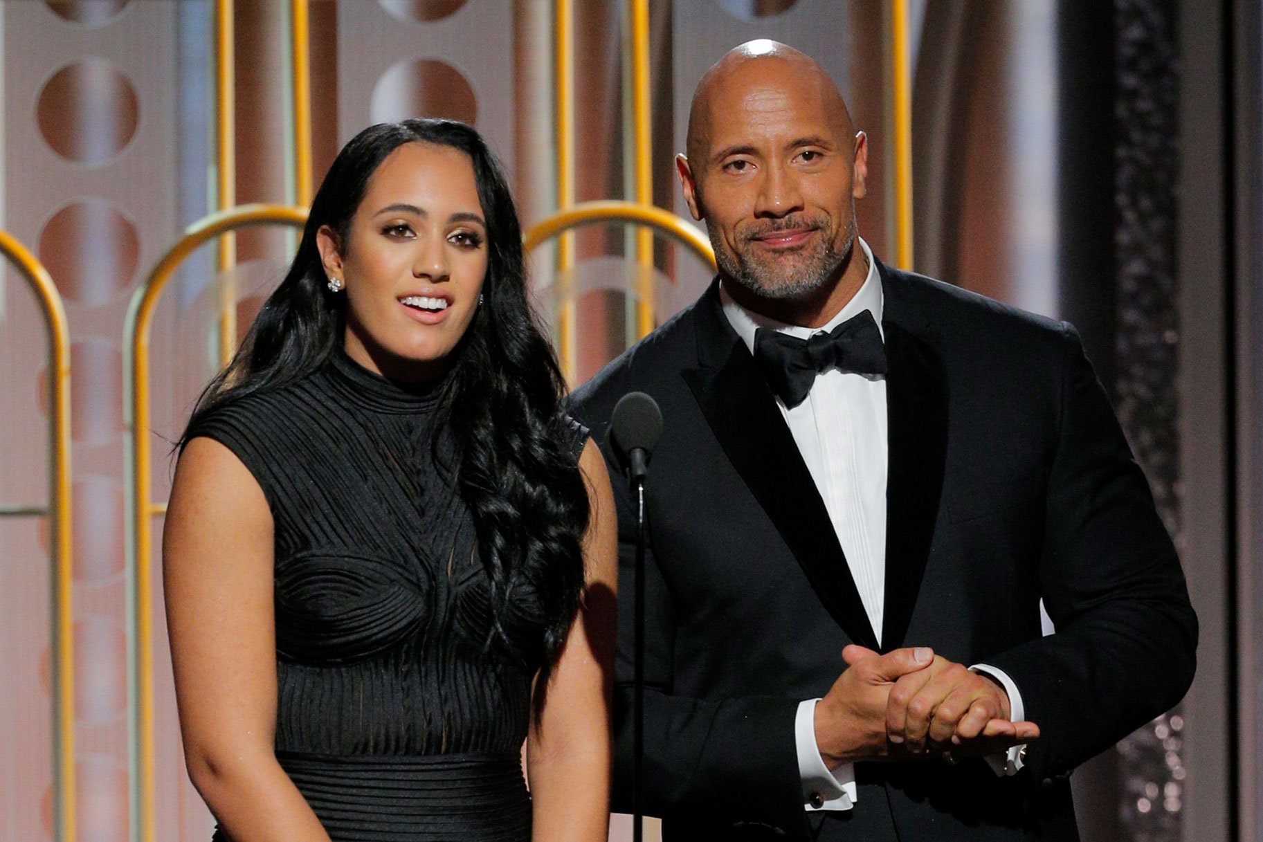 Simone Johnson and The Rock dressed in formal, black attire, standing in front of a mic