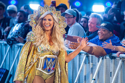 Charlotte Flair walking into the WWE ring wearing an extravagant gold jacket and headpiece. She is smiling